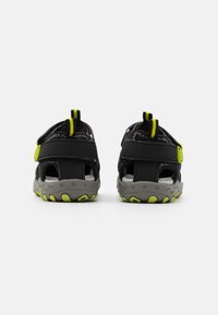 Sandalias negras de espalda abierta con suelas grises y acentos en verde neón. Cuentan con correas ajustables y una suela de goma texturizada.