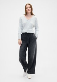 Svetlo sivi pulover z v-izrezom in dolgimi rokavi, kombiniran z ohlapnimi temno sivimi hlačami z elastičnim pasom. Model nosi bele superge.