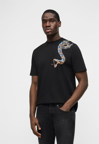 Άνδρας που φοράει μαύρο T-shirt με μεγάλο πολύχρωμο γραφικό φιδιού στο πάνω αριστερό μέρος του στήθους, συνδυασμένο με μαύρο τζιν, σε ουδέτερη στάση.