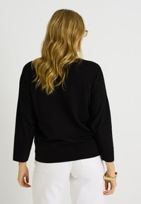 Suéter negro de corte amplio con mangas largas y anchas. La parte trasera tiene un dobladillo redondeado, combinado con pantalones blancos y una pulsera dorada.