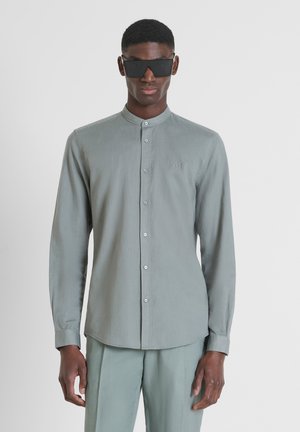Maglietta a maniche lunghe grigia chiara con colletto a band, bottoni frontali e ricamo del logo discreto. Indossata con pantaloni grigi chiari abbinati e occhiali da sole.