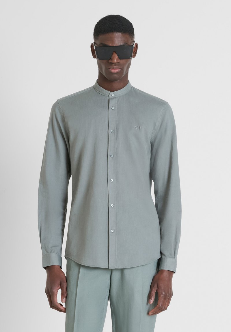 Maglietta a maniche lunghe grigia chiara con colletto a band, bottoni frontali e ricamo del logo discreto. Indossata con pantaloni grigi chiari abbinati e occhiali da sole.