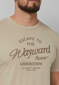 Beige katoenen t-shirt met bruin bedrukte tekst "ESCAPE TO THE Wayward Spirit" met een ronde halslijn en korte mouwen.