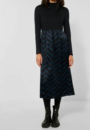 Kobieta ubrana w czarną sukienkę z długim rękawem, niebieską spódnicę w wzór chevron, czarne legginsy i czarne masywne buty.