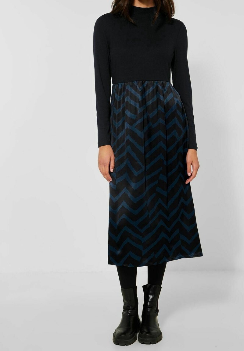 Femme portant une robe noire à manches longues avec une jupe bleue à motif chevron, des leggings noirs et des bottes épaisses noires.