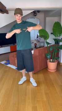 Zelené tričko s logom, čierne denimové šortky a biele tenisky. Interiér s hnedou pohovkou a veľkými zelenými rastlinami. Drevená podlaha.