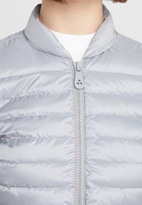 Ljusgrå puffjacka med ståkrage, horisontell sömnad och dragkedja framtill. Materialet verkar slätt och något glänsande.