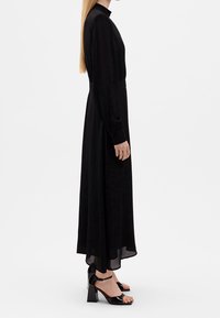 Langes schwarzes Kleid mit hohem Ausschnitt, langen Ärmeln und taillierter Taille. Transparenter unterer Saum und elegante, glänzende schwarze Blockabsatzsandalen.