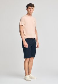 Lichtroze t-shirt en marineblauwe shorts op een mannelijke model. Het t-shirt heeft een ronde hals en korte mouwen, met een klein embleem op de borst.