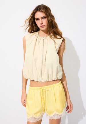 Femme aux longs cheveux portant un haut beige sans manches avec fermeture éclair devant et un short jaune à cordon avec bordure en dentelle blanche, debout sur fond blanc.