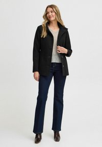 Zwarte wollen jas met een reverskraag, knopen aan de voorkant en twee zijzakken, gecombineerd met een grijze trui en donkere denim jeans.