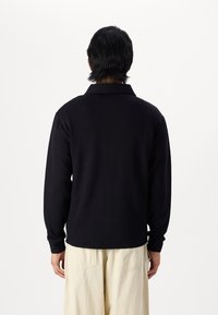 Polo noir à manches longues avec col, texture lisse et coupe légèrement décontractée, associé à un pantalon beige clair visible en dessous.