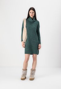Knit, teal dress with a high collar and side slit, paired with cream fur-accented boots. Features a soft texture and relaxed fit.