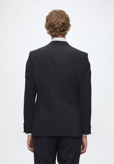 Homme aux cheveux blonds bouclés portant un costume noir et une chemise blanche, debout de dos sur un fond blanc uni.