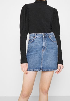 Femme portant un haut texturé noir à manches longues avec des épaules bouffantes et un col montant, associé à une mini-jupe en jean bleue.