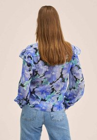 Blouse fleurie en tissu transparent, présentant des motifs bleus et verts sur un fond noir, avec des détails froncés aux manches et aux poignets.