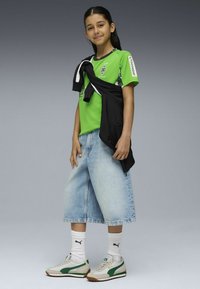 Zielona koszulka sportowa, czarna kurtka zarzucona, luźne szorty dżinsowe i białe skarpetki z logo. Obuwie: szare trampki z zielonymi akcentami.