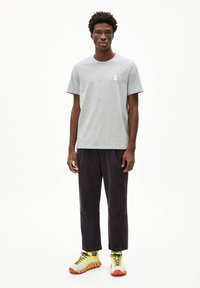 Grijze katoenen T-shirt met korte mouwen en een klein wit lintembleem, gecombineerd met donker corduroy broeken en heldere sneakers met gele en oranje accenten.
