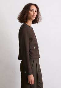 Braune Wildlederjacke mit Rundhalsausschnitt, langen Ärmeln, aufgesetzten Fronttaschen und silbernen Druckknöpfen, präsentiert an einem Modell vor einem hellen Hintergrund.