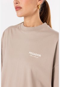 Bežové oversize tričko s klasickým okrúhlym výstrihom a vyšitým bielym logom "PEGADOR" na ľavej hrudi. Mäkko, bavlnené materiál.