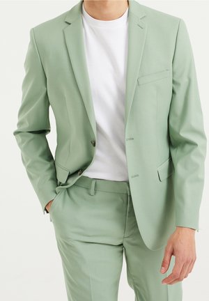 Mężczyzna w jasnorozowym garniturze i białym t-shircie, jedna ręka w kieszeni, na tle jednolitego jasnego tła.