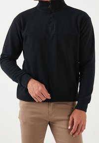 Pullover in pile nero con colletto alto, dotato di mezza zip, maniche lunghe e texture liscia. Abbinato a pantaloni beige.