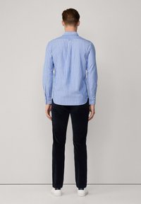 Camisa de manga larga azul claro con rayas blancas verticales, puños abotonados y detalle de pliegues en la parte posterior, combinada con pantalones oscuros y zapatillas blancas.