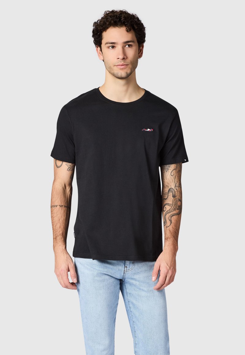 Ung mand med krøllet hår iført en sort t-shirt med lille logo på brystet og lyseblå jeans, stående foran en ensfarvet baggrund.
