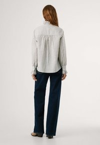 Svetlo siva karirana srajca z visokim ovratnikom, dolgimi rokavi in nabranim hrbtom. Obuta z temno modrimi širokimi hlačami in bež petami.