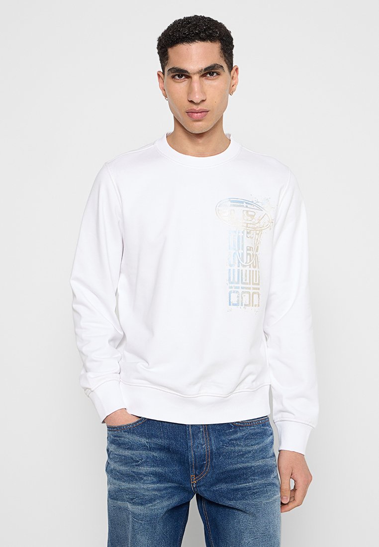 Giovane uomo con capelli corti e ricci, che indossa un felpa bianca con una grafica verticale sbiadita e jeans blu, in piedi con una mano nella tasca.