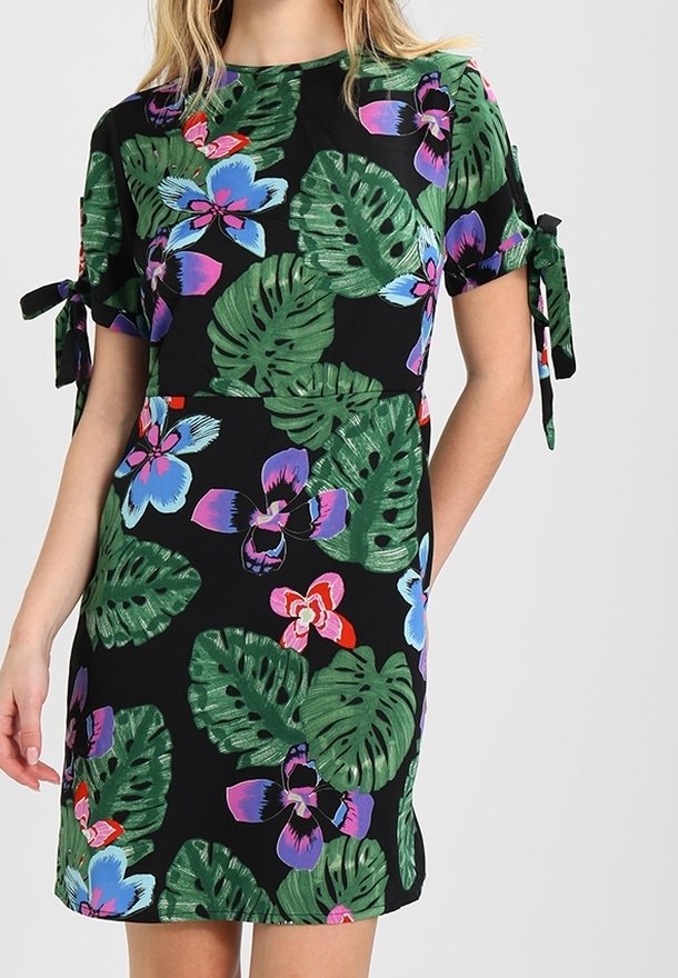 Femme portant une courte robe noire avec des feuilles tropicales vertes, des fleurs bleues, violettes et rouges, et des nœuds noués sur les manches courtes.