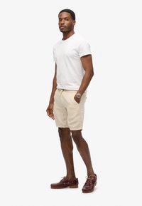 Camiseta blanca de cuello redondo, pantalones cortos beige con cordón y zapatos de barco marrones. Reloj notable en la muñeca. Diseño simple, estilo casual, colores neutros.