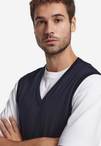 Mörkblå stickad väst med V-ringning, bärs över en vit T-shirt, med mjuk textur och avslappnad passform. Armar korsade framför kroppen.