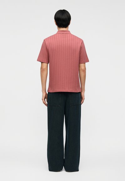 Άτομο με κοντά μαύρα μαλλιά, που φοράει ριγωτό ροζ κοντομάνικο πουκάμισο και φαρδιά μαύρη παντελόνια, στέκεται γυρισμένο προς τα πίσω σε λευκό φόντο.