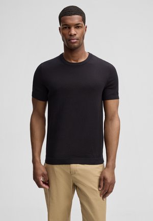 Mann, der mit dem Gesicht nach vorne steht und ein schwarzes Kurzarm-Pulloverhemd und beige Hosen vor einem schlichten grauen Hintergrund trägt.