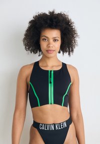 Čierny športový top s krátkym strihom, zelenými akcentmi a predným zipsom, v kombinácii s čiernymi bikiny nohavičkami s bielym pásom "CALVIN KLEIN".