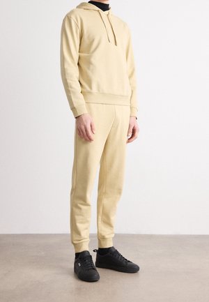 Mand iført beige hættetrøje og matchende joggingbukser med sorte sneakers, der står på gråt gulv mod hvid væg.