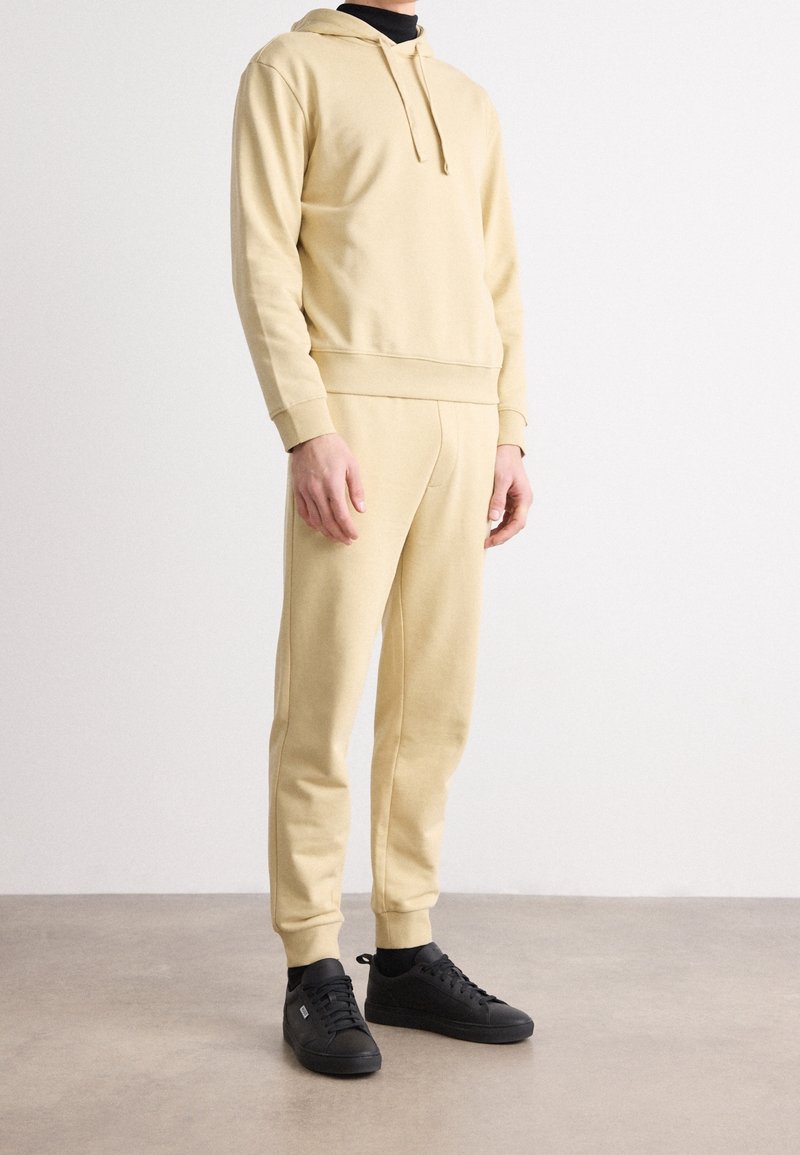Homme portant un sweat à capuche beige et un pantalon de jogging assorti avec des baskets noires, debout sur un sol gris contre un mur blanc.