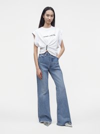 Jeune femme portant un top sans manches blanc Marc Jacobs, un jean taille haute à jambes larges et des chaussures argentées pointues, debout avec une main dans la poche.