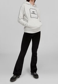 Persona che indossa un cappuccio O'Neill grigio chiaro, pantaloni neri svasati e scarpe da trekking nere, in piedi con le mani nelle tasche del cappuccio.