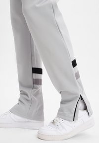 Helle graue Sporthosen mit Reißverschlussbund, die an den Seiten schwarze und graue Streifen aufweisen. Getragen mit weißen Sneakers und strukturiertem Stoff.