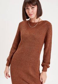 Vestido de suéter marrón de punto con mangas largas, textura acanalada, escote en forma de V y silueta ajustada. Lleva puesta una sencilla cadena.
