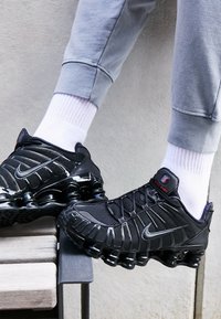Τα μαύρα αθλητικά παπούτσια Nike Shox διαθέτουν κομψό σχεδιασμό με λαμπερά στοιχεία, υφασμάτινο επάνω μέρος με υφή και εμφανή απορρόφηση κραδασμών στην σόλα, συνδυασμένα με λευκές κάλτσες.