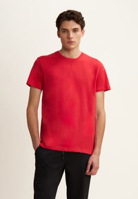 T-shirt in cotone rosso a maniche corte e collo rotondo, con texture liscia e design minimalista. Indossato con pantaloni neri, in piedi su uno sfondo neutro.