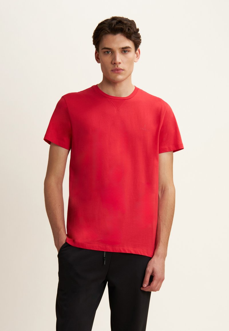 T-shirt in cotone rosso a maniche corte e collo rotondo, con texture liscia e design minimalista. Indossato con pantaloni neri, in piedi su uno sfondo neutro.