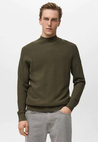 Maglione lavorato a maglia verde oliva con collo alto, polsini e orlo rigati. Presenta un motivo tessurizzato, abbinato a jeans grigio chiaro.