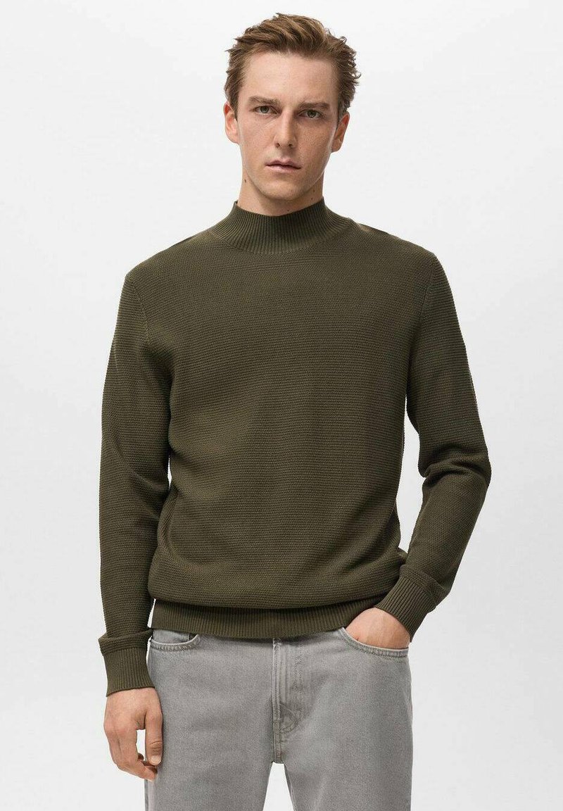 Maglione lavorato a maglia verde oliva con collo alto, polsini e orlo rigati. Presenta un motivo tessurizzato, abbinato a jeans grigio chiaro.