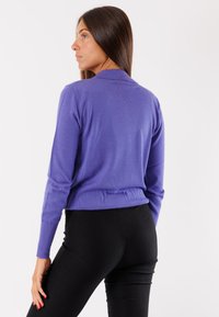 Maglione lungo a maniche con collare di colore viola, con polsini a coste. Indossato con pantaloni neri aderenti, che mostrano un fit rilassato e una texture morbida.