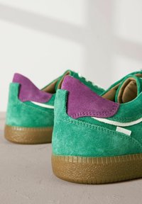 Baskets en velours vert avec un accent en velours violet au niveau du talon et une semelle en caoutchouc brun texturé. Elles présentent des coutures blanches et un motif de design subtil.