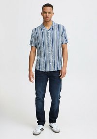 Majica s kratkimi rokavi in gumbi z modrimi in belimi navpičnimi črtami, sproščen kroj. V kombinaciji s temnimi jeans hlačami in belimi supergami.
